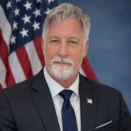 older man with white hair and beard in a suite with american flag in background