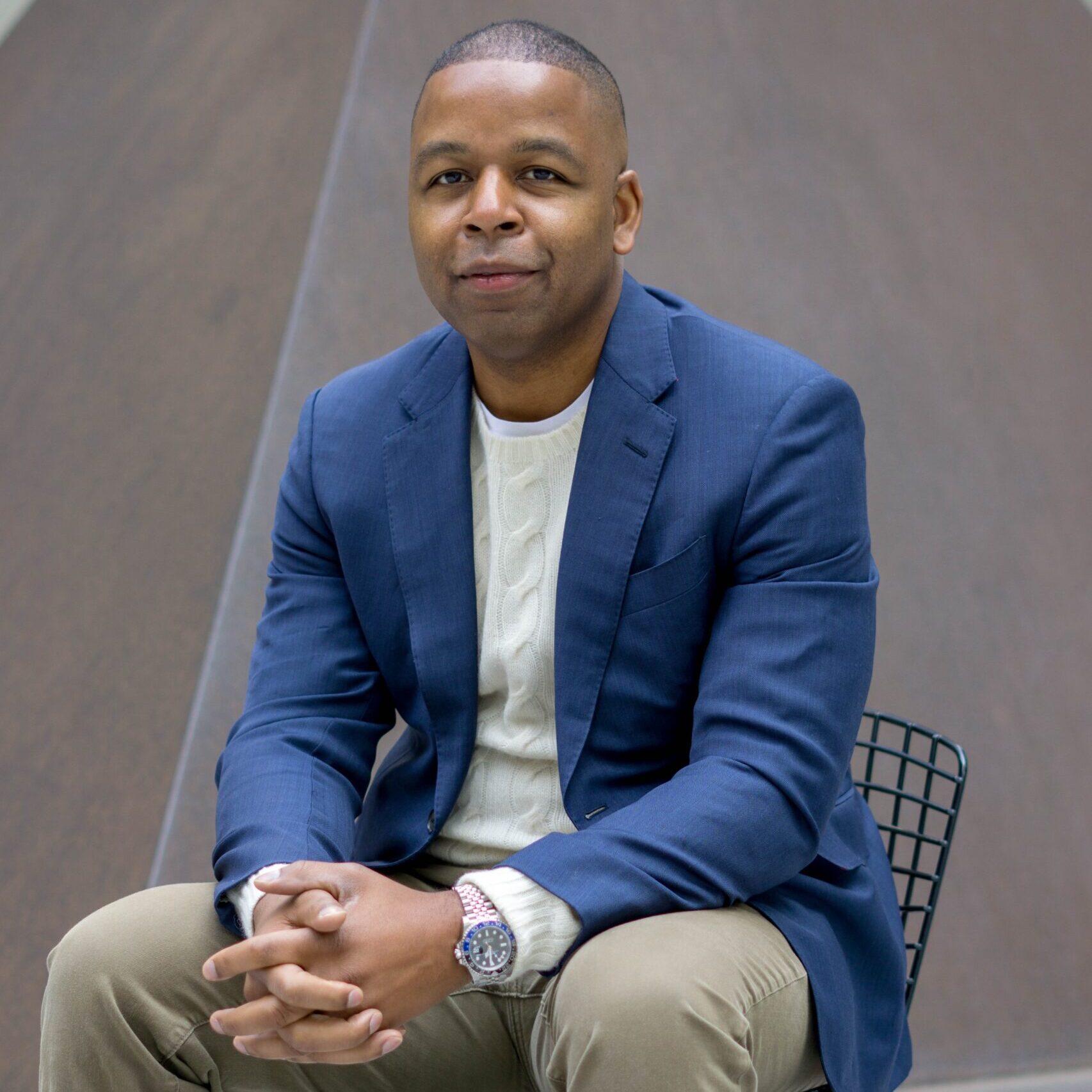 Man with dark skin in blue blazer and khaki pants sitting in a chair.